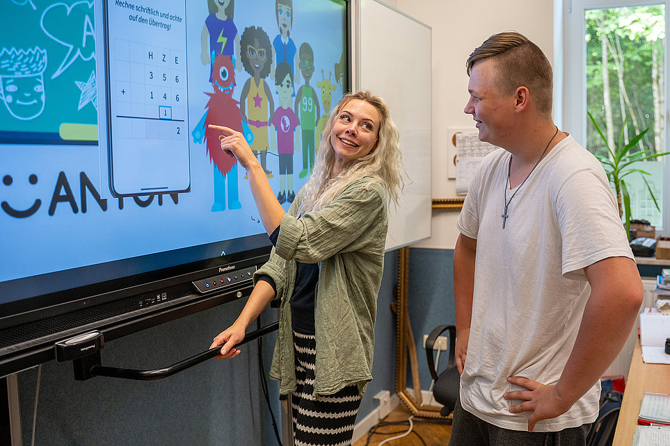 Lehrerin zeigt an großem Touchscreen ein Lernspiel; ein Schüler steht daneben, lächelt und beobachtet.