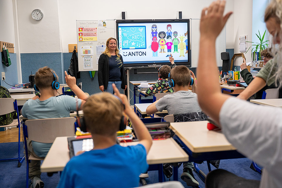 Lehrerin vor interaktivem Display, Schüler sitzen an Tischen mit Tablets, einige melden sich und tragen Kopfhörer — lebendiger Unterricht.