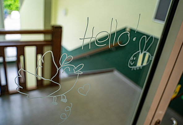 Auf Glasfenster gezeichnetes „Hello!“ mit Vogel‑ und Herzskizzen; verschwommene Treppe und Geländer im Hintergrund.