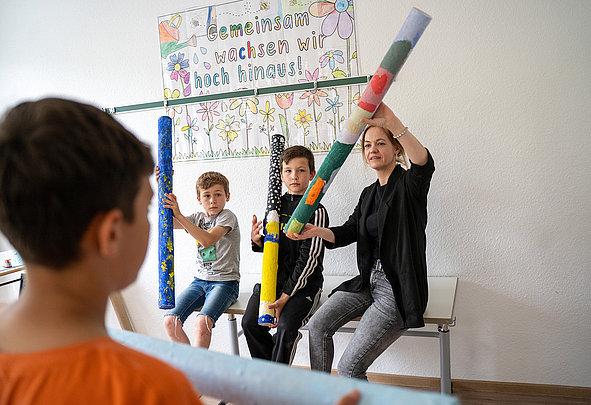 Drei Kinder und eine Betreuerin sitzen, halten bunte Schaumstoffrollen; Poster „Gemeinsam wachsen wir hoch hinaus!“ an der Wand.