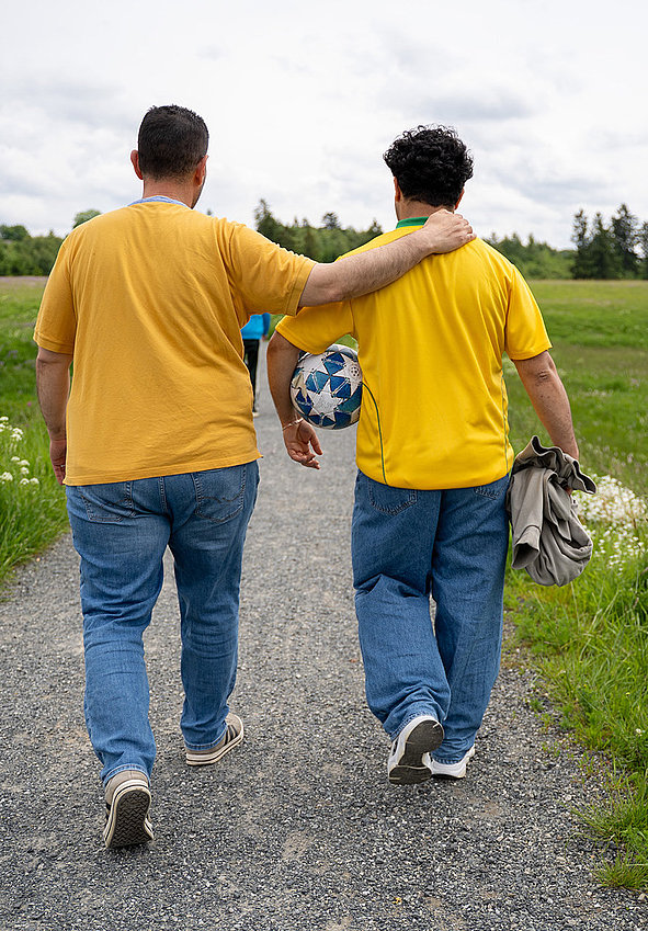 Zwei junge Männer in gelben Shirts gehen Arm in Arm auf einem Kiesweg durch grüne Wiesen; einer trägt einen Fußball, entspannte Stimmung.